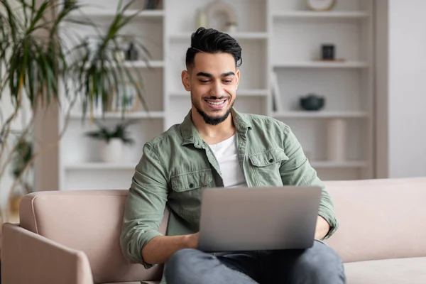 Feliz atractivo milenario árabe macho con barba en casual se sienta en el sofá, escribiendo en el ordenador portátil en la sala de estar — Foto de Stock