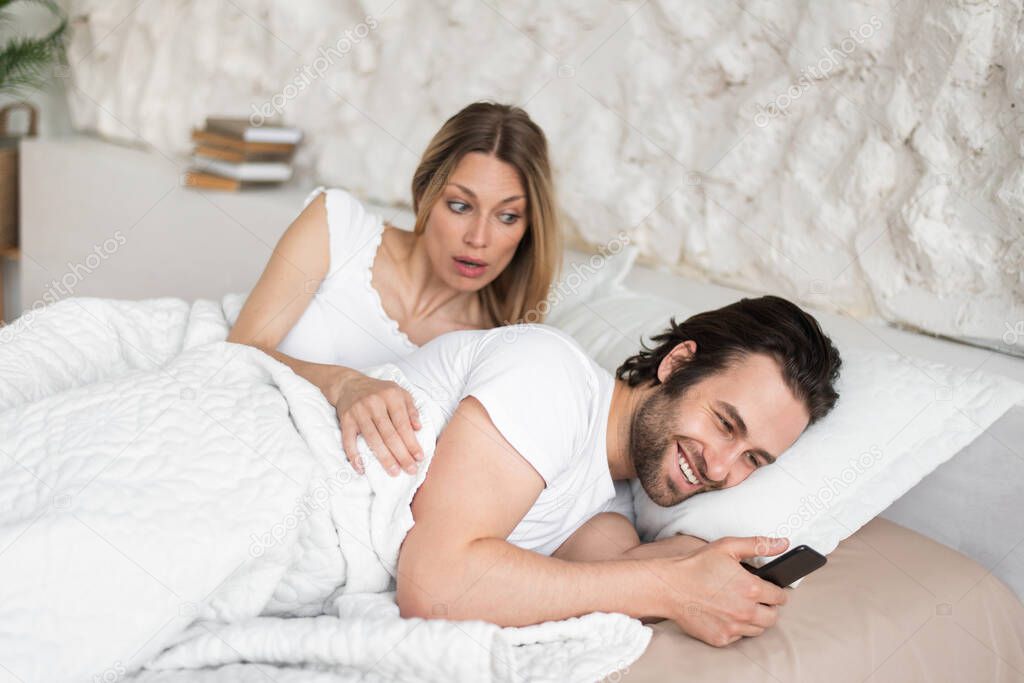 Shocked young woman seeing her husband messaging to his lover on smartphone while lying in bed at home
