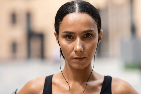 Confiado milenario árabe mujer atleta morena en los auriculares mira a la cámara en la construcción de fondo —  Fotos de Stock