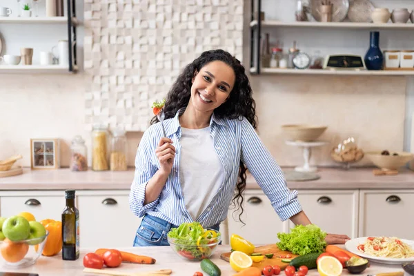 Feliz señora latina comiendo ensalada de verduras frescas, disfrutando de un almuerzo fresco, de pie en la cocina y sonriendo a la cámara — Foto de Stock