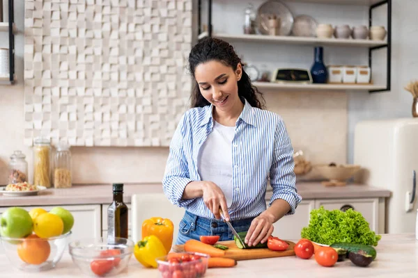 Joven latina cocinando deliciosa y saludable ensalada de pie en la cocina moderna, rebanando pepino en la tabla de cortar — Foto de Stock