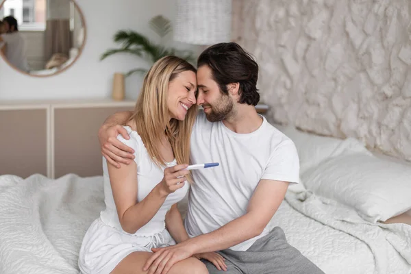 Mujer joven y feliz abrazando a su marido, con la prueba de embarazo positiva en la cama en casa — Foto de Stock