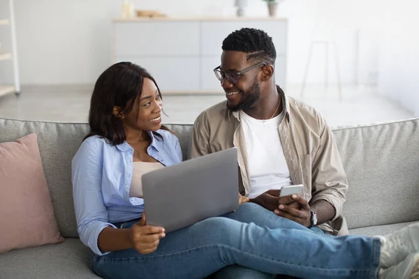 Happy black couple sitting on couch, using smartphone and pc
