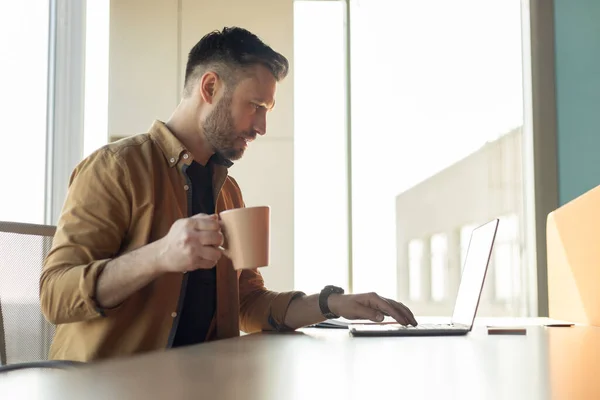 Mann arbeitet am Laptop und trinkt Kaffee im Büro, Seitenansicht — Stockfoto