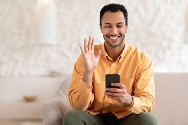 Gülümseyen genç adam cep telefonu kullanarak el sallıyor.