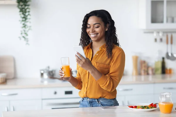 Positivo bonita dama negra sosteniendo vaso de jugo y teléfono inteligente —  Fotos de Stock