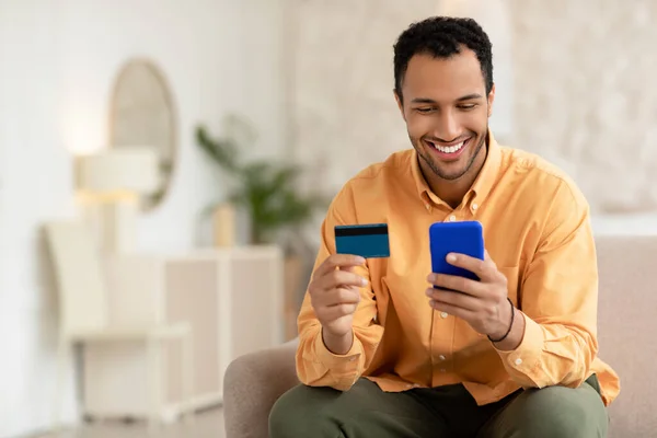 Lachende jonge man met behulp van mobiele telefoon en creditcard thuis — Stockfoto