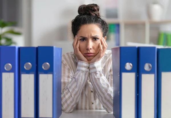 Upset lady businesswoman sitting at workplace with stack of folders and looking at camera, feeling tired — стоковое фото