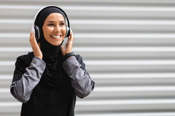 Wanita islamik milenial yang tersenyum dalam jilbab mengenakan headphone nirkabel melihat ruang kosong — Stok Foto