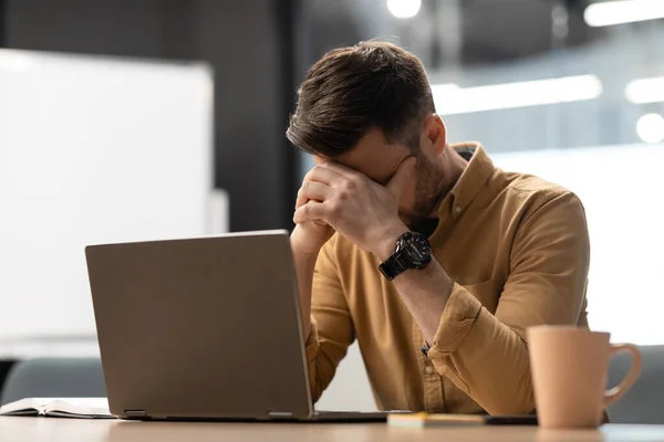 Verzweifelter Geschäftsmann in der Nähe eines Laptops, der Gesicht bedeckt und Probleme im Büro hat — Stockfoto