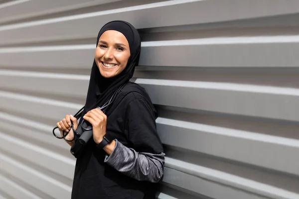 Tersenyum muda timur tengah atlet perempuan dalam jilbab dengan melompat-lompat tali dekat dinding abu-abu latar belakang, ruang kosong — Stok Foto
