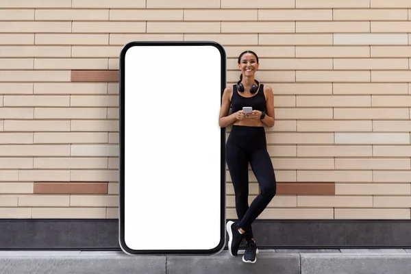 Joven atleta árabe sonriente en el puesto de ropa deportiva cerca de un enorme teléfono inteligente con pantalla en blanco —  Fotos de Stock