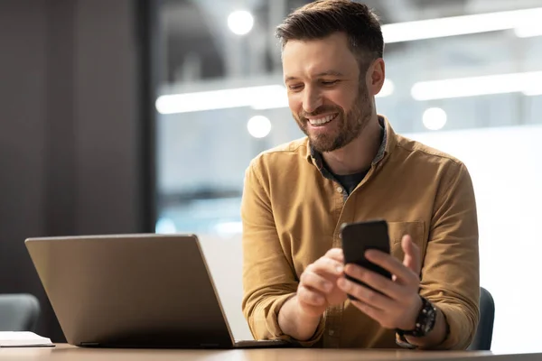 Feliz empresario hombre mensajes de texto usando Smartphone cerca de la computadora portátil en la oficina — Foto de Stock