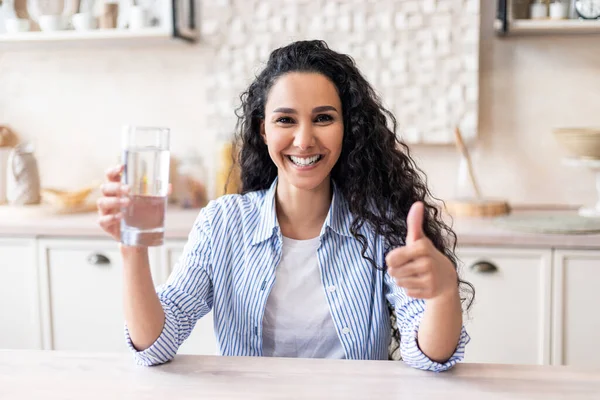 Vzrušená latinská žena držící sklo s čistou vodou a ukazující palec nahoru, sedící u stolu v kuchyni — Stock fotografie