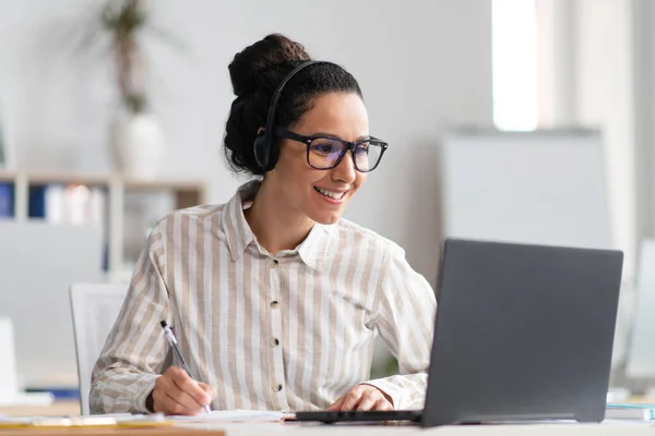 Porträtt av glad ung latinamerikansk kvinna bär trådlösa hörlurar titta på online kurs, sitter vid bordet — Stockfoto