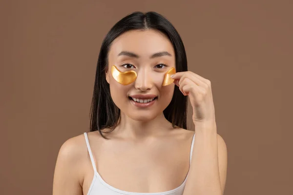 Retrato de mujer asiática joven quitando parches de ojo de hidrogel dorado sobre fondo de estudio marrón, sonriendo a la cámara — Foto de Stock