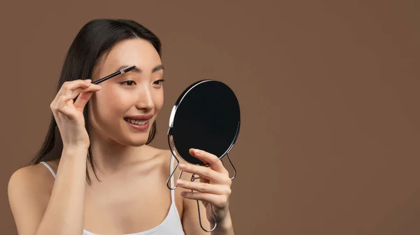 Naturlig makeup koncept. Glad asiatisk dam borsta ögonbryn, hålla borste och spegel, brun bakgrund — Stockfoto