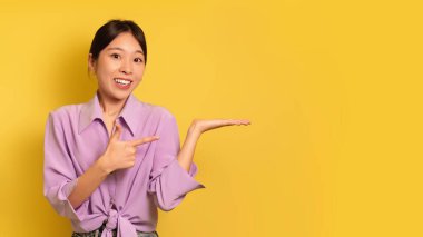 Neşeli genç Asyalı bayan avucundaki görünmez nesneyi sarı stüdyo arka planında gösteriyor.