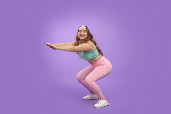 Happy young pretty european plus size female in sportswear squats, isolated on purple background — Stock Photo, Image