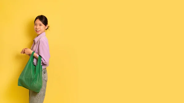 Pozitivní mladá asijská fena drží opakovaně použitelné eko textilní nákupní taška na žlutém pozadí studia, volné místo — Stock fotografie