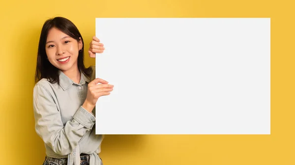 Encantadora joven asiática sosteniendo un póster de papel en blanco con maqueta, sonriendo a la cámara sobre fondo amarillo del estudio —  Fotos de Stock