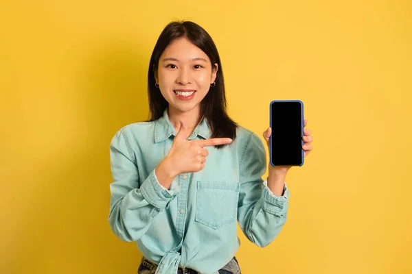 Lächelnde Asiatin zeigt auf Handy mit leerem Bildschirm, wirbt für Ihre Website, bietet Platz für Werbung, Attrappe — Stockfoto