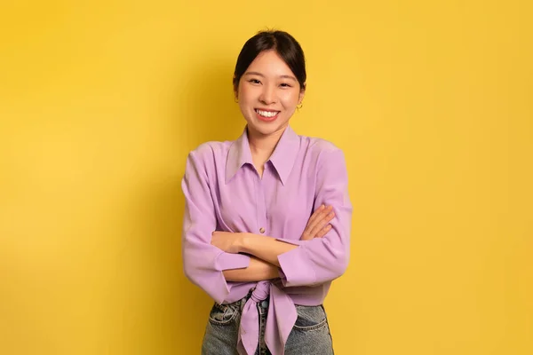 Positivo jovem mulher asiática de pé com braços cruzados e olhando para a câmera sobre o fundo do estúdio amarelo — Fotografia de Stock