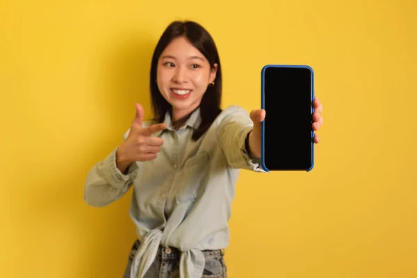 Fröhliche junge Asiatin zeigt auf Handy mit leeren Platz für mobile App oder Website auf dem Bildschirm, Attrappe — Stockfoto