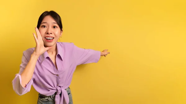 Izgalmas Y-generációs ázsiai nő kiabál kéz a száj közelében, és rámutatva az üres tér sárga stúdió háttér — Stock Fotó