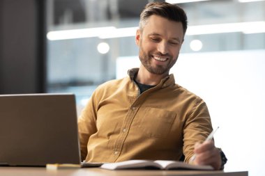 Ofiste dizüstü bilgisayarla çalışan mutlu iş adamı not alıyor.