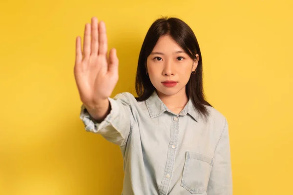 Fiatal ázsiai hölgy gesztikulál STOP, mutató tenyér kamera sárga stúdió háttér — Stock Fotó