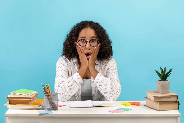 Chockad ung svart kvinna i glasögon sitter vid bordet med studiematerial på blå studio bakgrund — Stockfoto