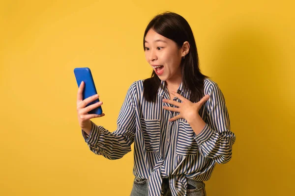 Shocked young Asian lady looking at smartphone screen, shouting in excitement, winning lottery on yellow background — 스톡 사진