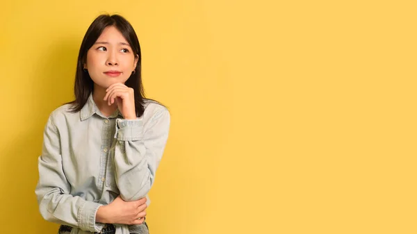 Pensativo jovem ásia mulher no casual roupa olhando para vazio espaço sobre amarelo estúdio fundo, panorama — Fotografia de Stock