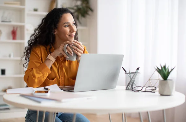 Mujer de negocios soñadora usando el ordenador portátil y sosteniendo la taza de café en el interior —  Fotos de Stock