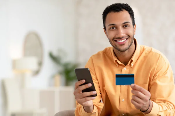 Joven sonriente usando smartphone y tarjeta de crédito en casa —  Fotos de Stock