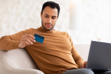 Gülümseyen adam evde bilgisayar ve kredi kartı kullanıyor.