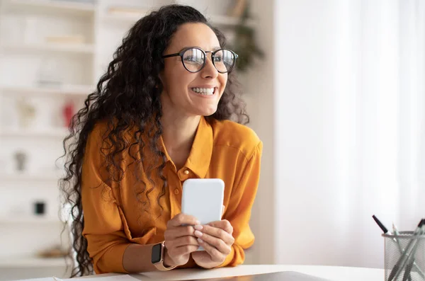 Sorridente signora araba utilizzando smartphone guardando oltre seduto sul posto di lavoro — Foto Stock