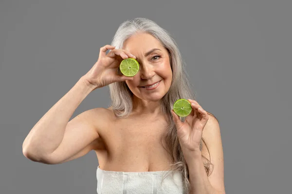 Vitaminas de fruto naturais de pele madura. Mulher sênior adorável com cabelo comprido segurando limão perto de seu rosto em fundo cinza — Fotografia de Stock