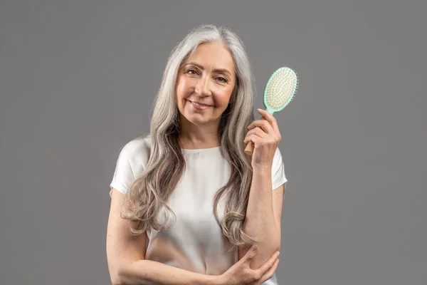 Portrait of lovely senior lady with beautiful long hair holding brush and smiling at camera on grey studio background — 스톡 사진