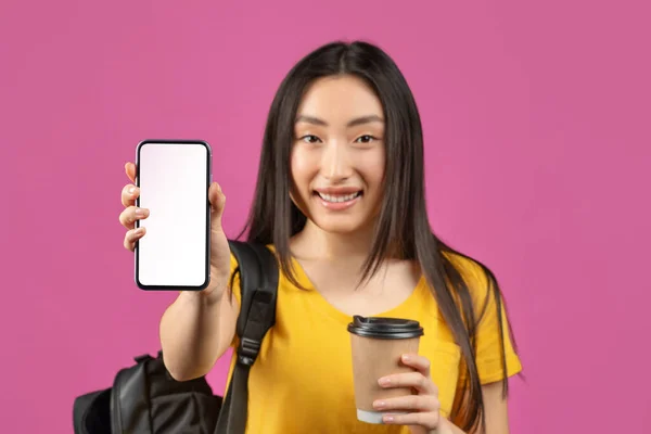 Positieve Aziatische vrouwelijke student toont smartphone met wit leeg scherm, poseren met rugzak en koffie, mockup — Stockfoto