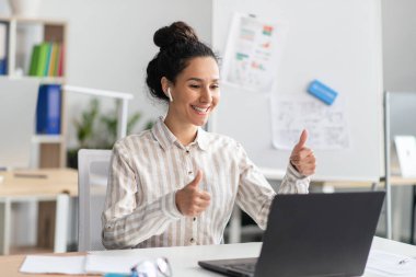 Mutlu CEO hanımın dizüstü bilgisayarla video konferansı var ve web kamerasında baş parmak işareti yapıyor, ofisinde oturuyor.