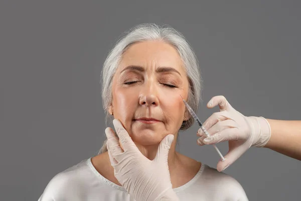 Concepto de inyecciones de belleza. Atractiva mujer mayor recibiendo ácido hialurónico o inyección de colágeno en fondo de estudio gris — Foto de Stock