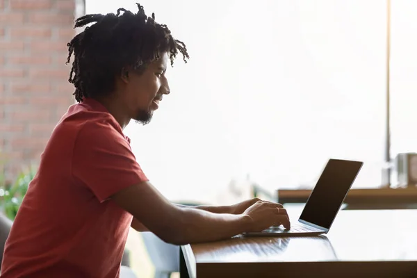 Boldog millenniumi fekete göndör férfi ül asztalnál, gépelés a számítógépen kávézó belső ablak háttér, napkitörés — Stock Fotó
