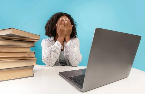 Jonge Afro-Amerikaanse studente die haar gezicht bedekt, zit achter een bureau met boeken, huilt voor het laptopscherm — Stockfoto