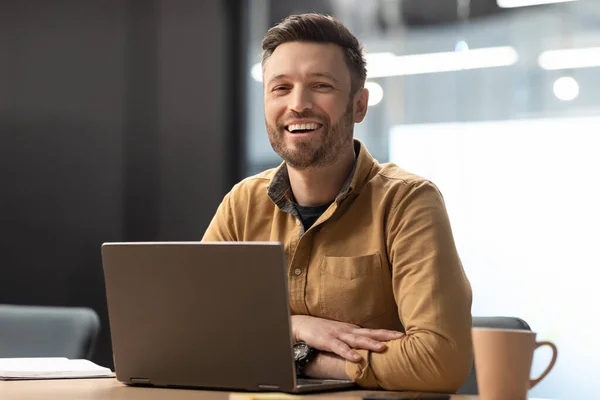 Alegre empresario masculino posando cerca del ordenador portátil que trabaja en la oficina moderna — Foto de Stock