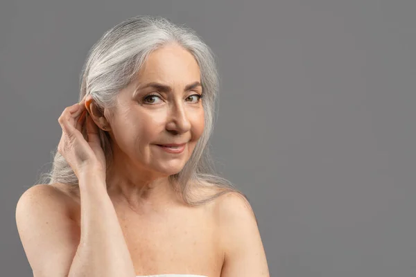 Kecses öregedés. Szépség portré idős nő beállítása hosszú haj a füle mögött szürke stúdió háttér, másolás tér — Stock Fotó