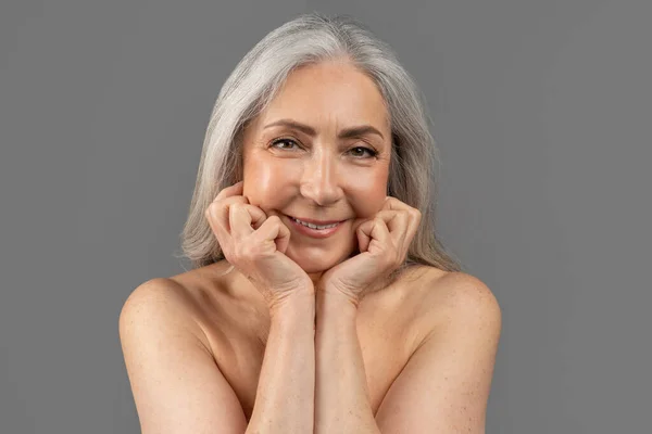 Beautiful mature woman with nude body looking at camera and smiling, holding hands near face on grey studio background — 스톡 사진