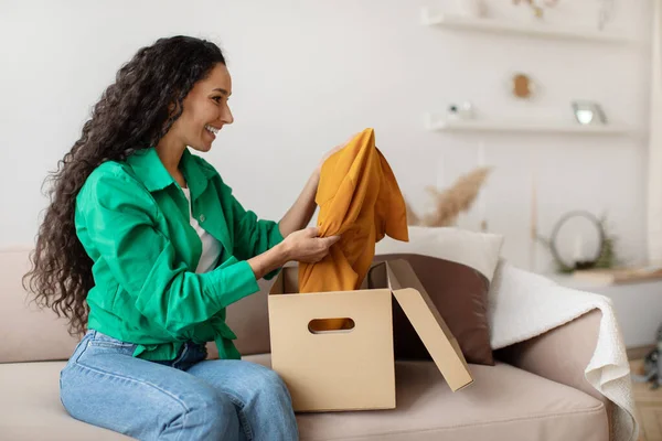Щаслива жінка тримає одяг розпаковуючи картонну коробку вдома — стокове фото