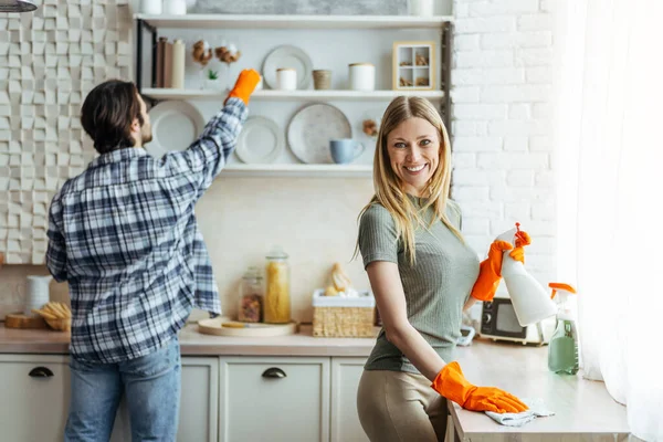 Happy young european female and male in rubber gloves with cleaning supplies in hands, clean dust in kitchen — Stock Photo, Image